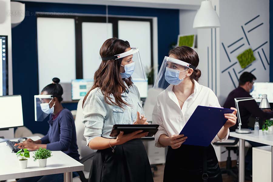 戴着防冠状病毒防护面罩的女企业家站在工作场所谈论公司财务数据，手持数字平板电脑。尊