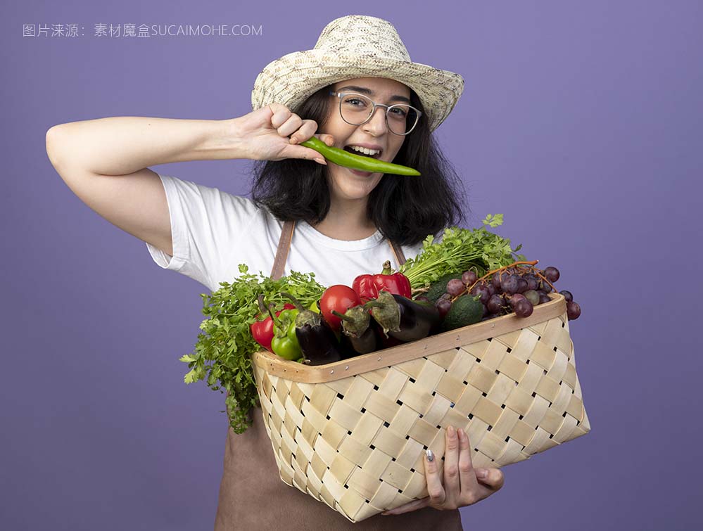 快乐年轻的黑发女性园丁在光学眼镜和统一戴园艺帽子拿着蔬菜篮子