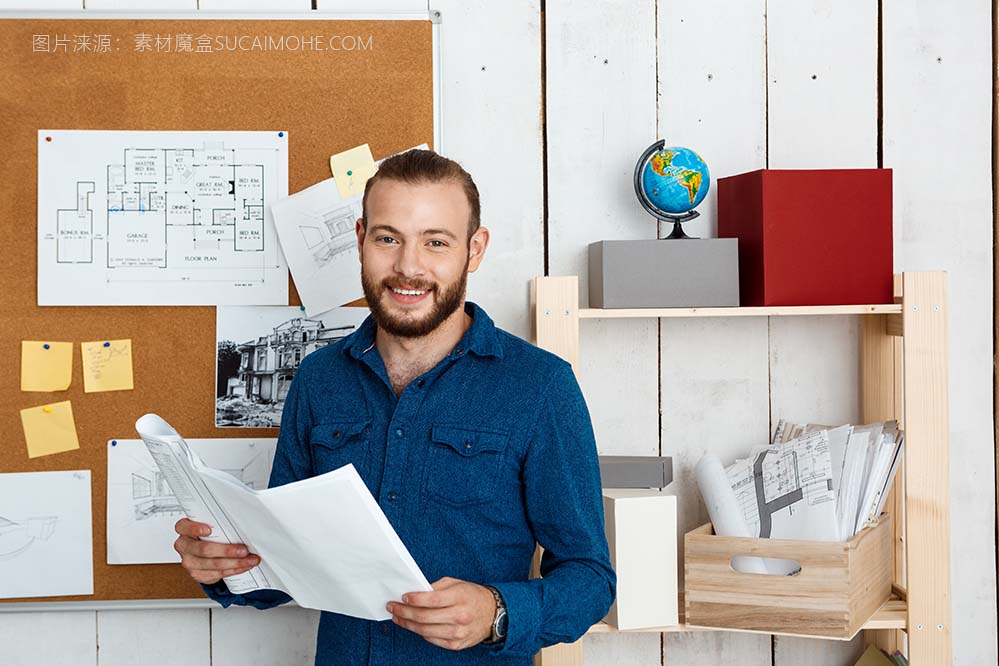年轻成功的建筑师微笑着拿着图纸站在办公室的墙上
